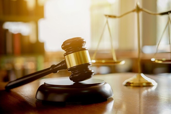 A wooden judge's gavel rests on its sound block with a blurred set of scales of justice in the background on a wooden desk.
