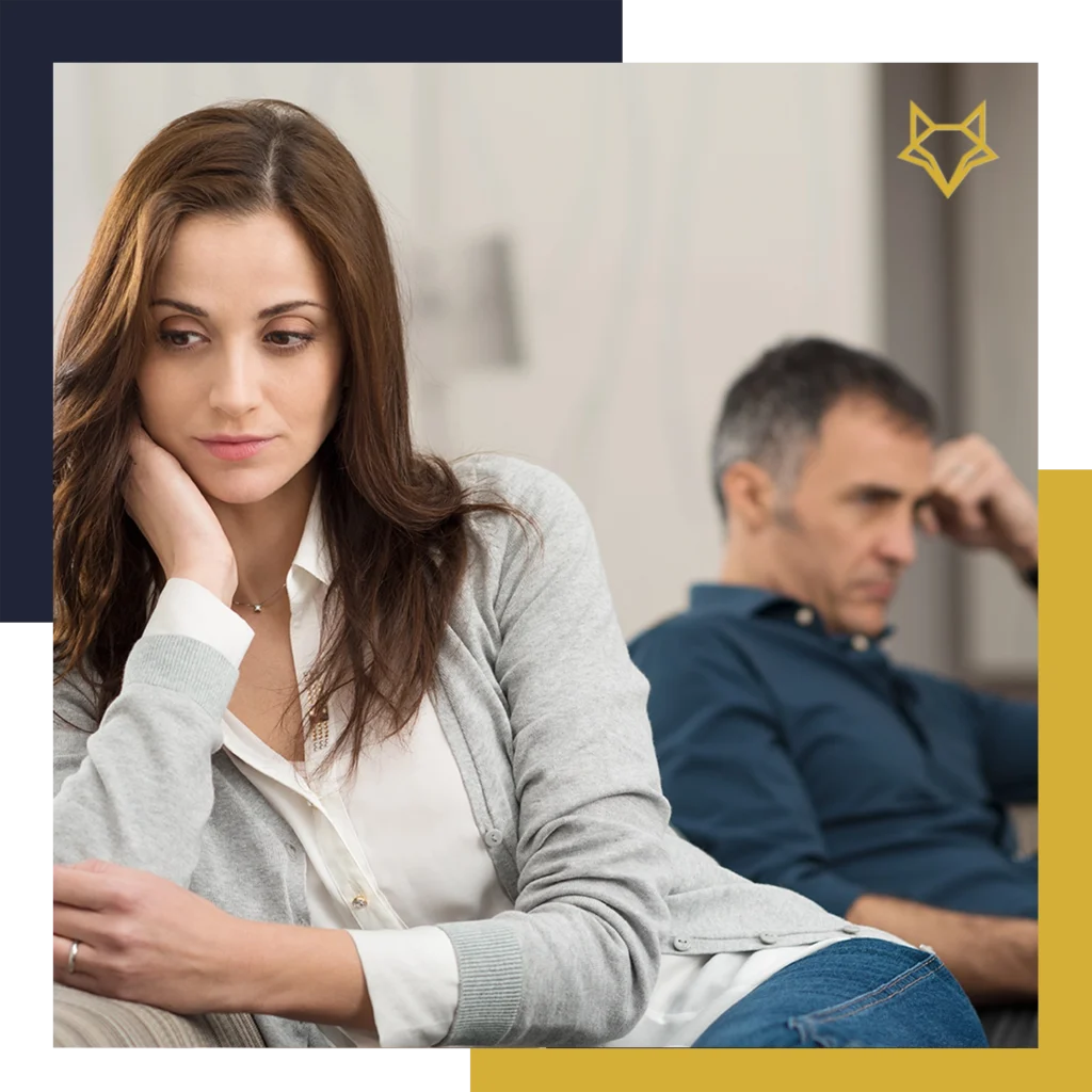 A woman sits in the foreground looking pensive while a man sits in the background appearing upset; both look away from each other.