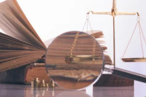 An open book, a brass balance scale, and a magnifying glass are arranged on a reflective surface, symbolizing law and investigation.