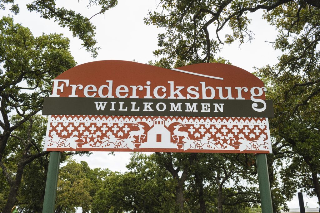 A sign reading "Fredericksburg Willkommen" stands among trees, featuring decorative patterns and a small church illustration.