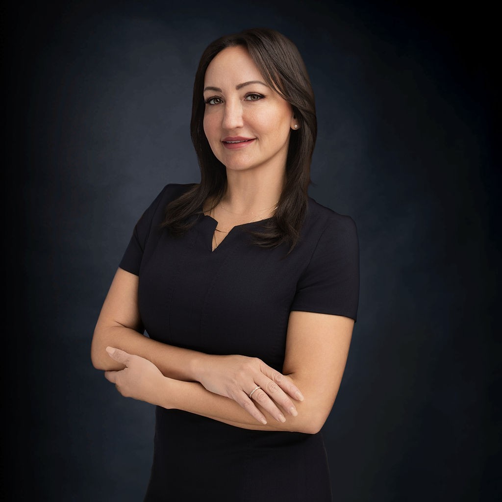 Woman with long brown hair wearing a black short-sleeve dress stands with arms crossed, looking at the camera against a dark background.