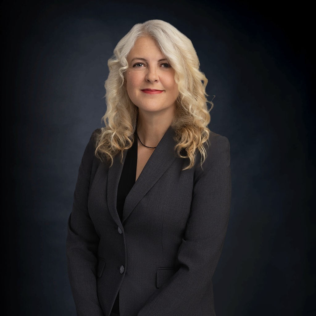 A woman with long, curly blonde hair wearing a dark suit jacket stands in front of a dark background, looking at the camera with a neutral expression.