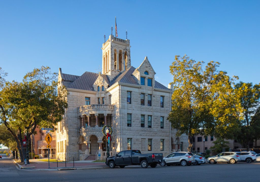 Historic stone courthouse building with a central tower, arched windows, and decorative wreath, surrounded by parked cars and trees under a clear blue sky.