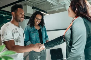 Two people stand by a desk, smiling as one shakes hands with a third person in business attire in an office setting.