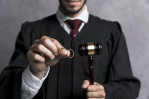 A person in a judge's robe holds a gavel in one hand and two wedding rings in the other, suggesting a legal decision about marriage or divorce.