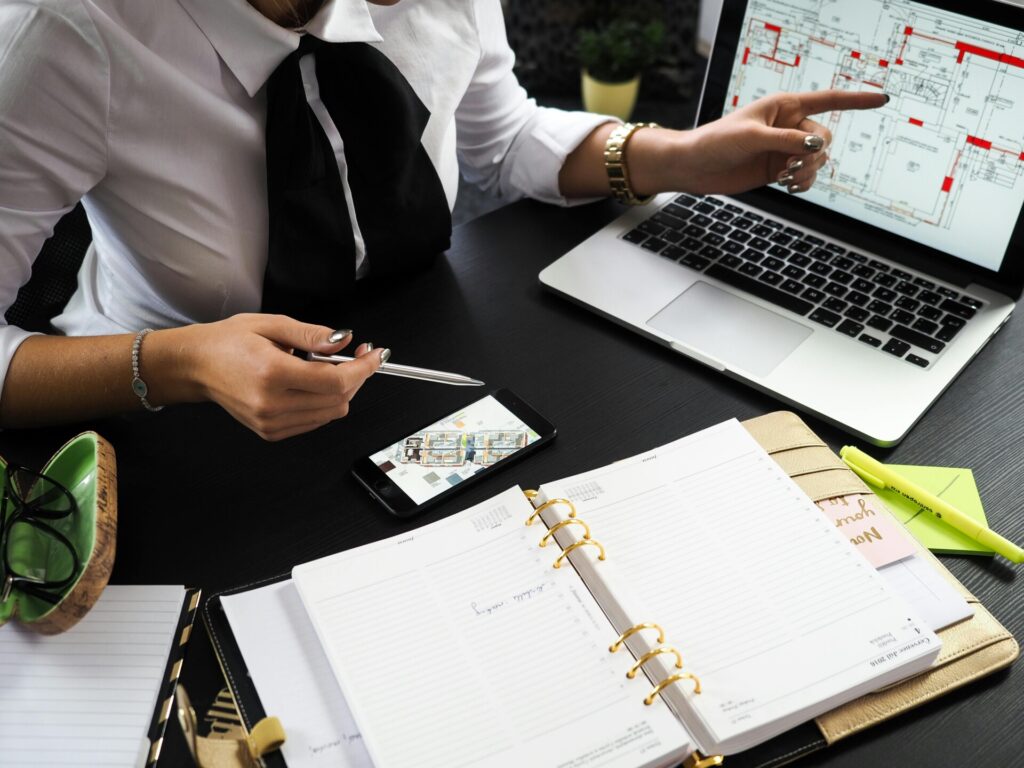 Person in business attire sits at a desk with an open planner, a smartphone, papers, and a laptop displaying architectural plans, pointing at the laptop screen.