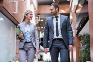 Two business professionals in suits walk and converse in a modern office corridor, one holding green folders.