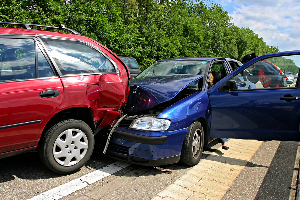 A blue car with a crumpled hood has rear-ended a red station wagon on a road, with both vehicles damaged and doors open.