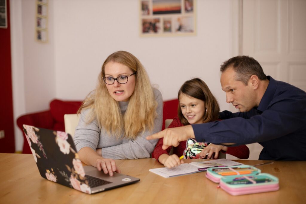 A woman, a young girl, and a man sit at a table, looking at a laptop together. The man is pointing at the screen and the girl is smiling. Papers and colored pencils are on the table.