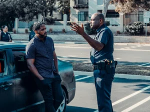 A police officer talks to a man leaning against a car in a parking lot, with another officer and person in the background.
