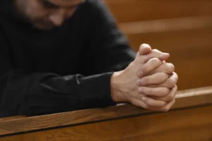 Person sitting in a wooden pew with hands clasped together, head bowed, suggesting prayer or contemplation.