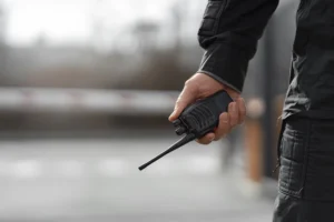 A person holding a black walkie-talkie outdoors, wearing a dark jacket, with a blurred background.