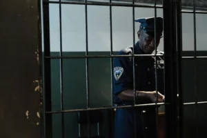 A police officer in uniform stands behind metal bars in a dimly lit room, appearing to unlock or secure the gate.