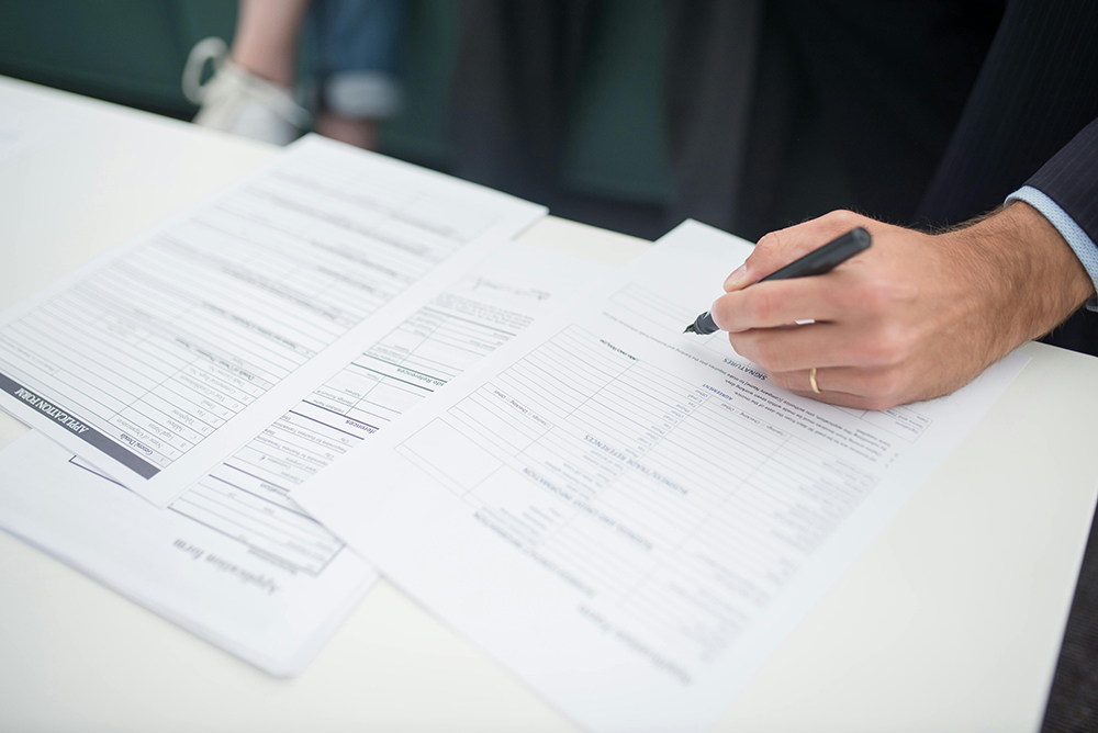 A person fills out paperwork on a white desk, holding a pen in their right hand, with several documents spread out in front of them.