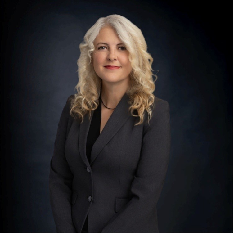 A woman with long, curly blonde hair wearing a dark blazer poses against a dark studio background.