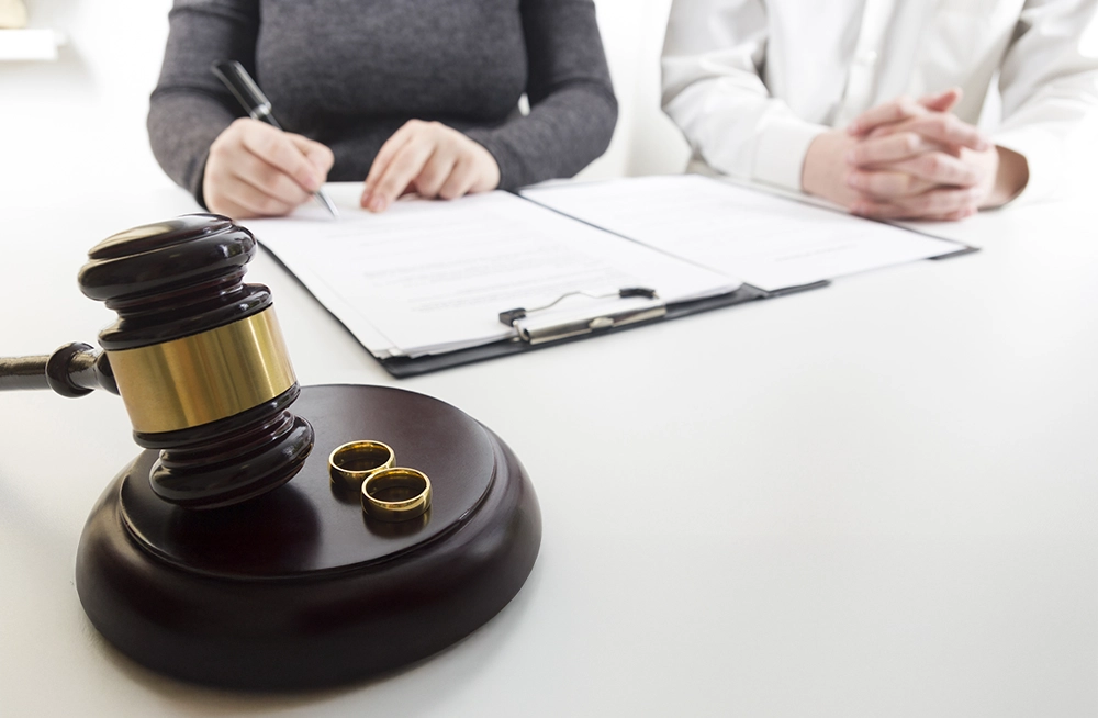 A gavel and two wedding rings are on a table, while two people sit nearby with legal documents, one writing and the other with hands clasped.