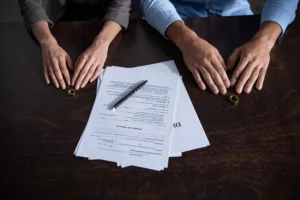Two people sit across from each other at a table with divorce papers, a pen, and wedding rings placed beside their hands.