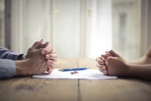 Two people sit across from each other at a table with hands clasped, a pen, and a document between them.