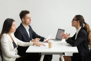 Three people in business attire are seated at a table having a discussion; two are on one side with documents and a laptop, while the third speaks from the other side.