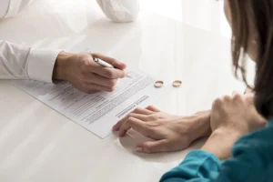 Two people sit at a table with a divorce agreement; one is signing the document while two wedding rings rest on the table.