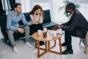 A couple sits on a couch speaking with a professional man across a coffee table with notebooks, hourglass, and mugs on it.