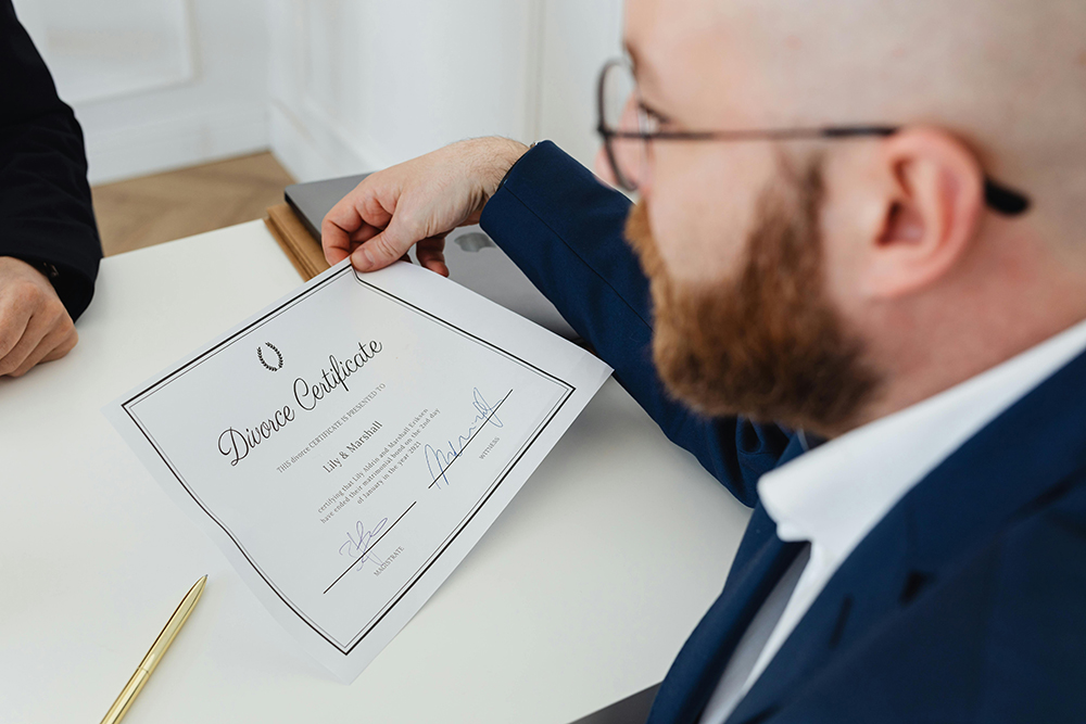 A man in a suit holds a divorce certificate while seated at a desk, with a gold pen and another person’s hand visible nearby.
