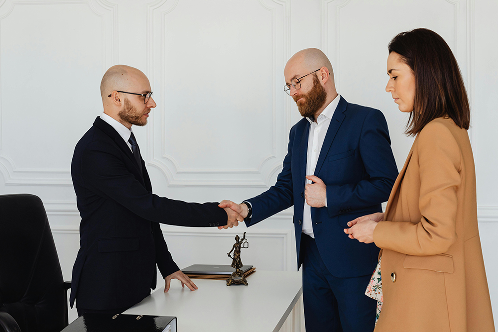 Two men in suits shake hands across a desk in an office, while a woman in a tan coat stands nearby observing.