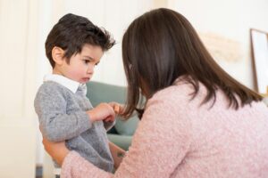 A woman crouches in front of a young boy, holding his arms and looking at him closely, while the boy looks down, appearing serious or upset.