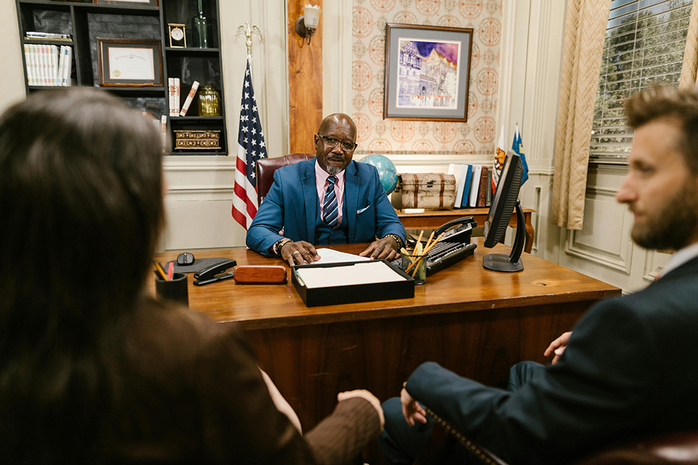 A man in a suit sits at his desk in an office, facing two people seated across from him. An American flag and framed picture are visible in the background.