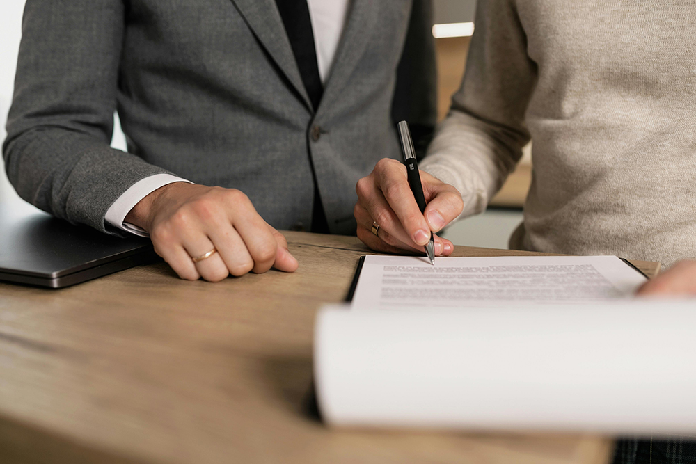 Two people stand at a desk; one in a suit observes while the other writes on a document with a pen. A closed laptop and a clipboard are also on the desk.