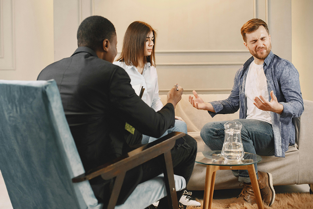 Three people sit in a conversation; a man gestures emotionally, while a woman and another man, possibly a counselor, listen attentively in a modern room.