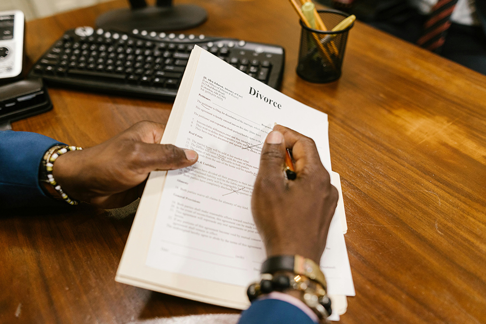 A person holding a pen signs a divorce document at a wooden desk, with a keyboard, pen holder, and papers visible in the background.