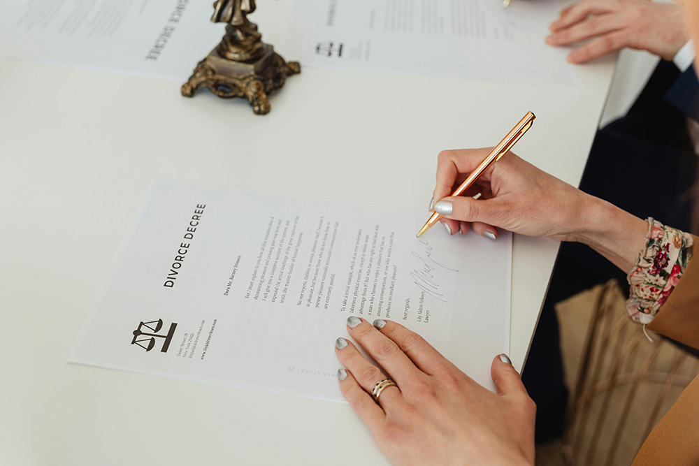 A person signs a divorce decree document at a table, with another person’s hands visible in the background.