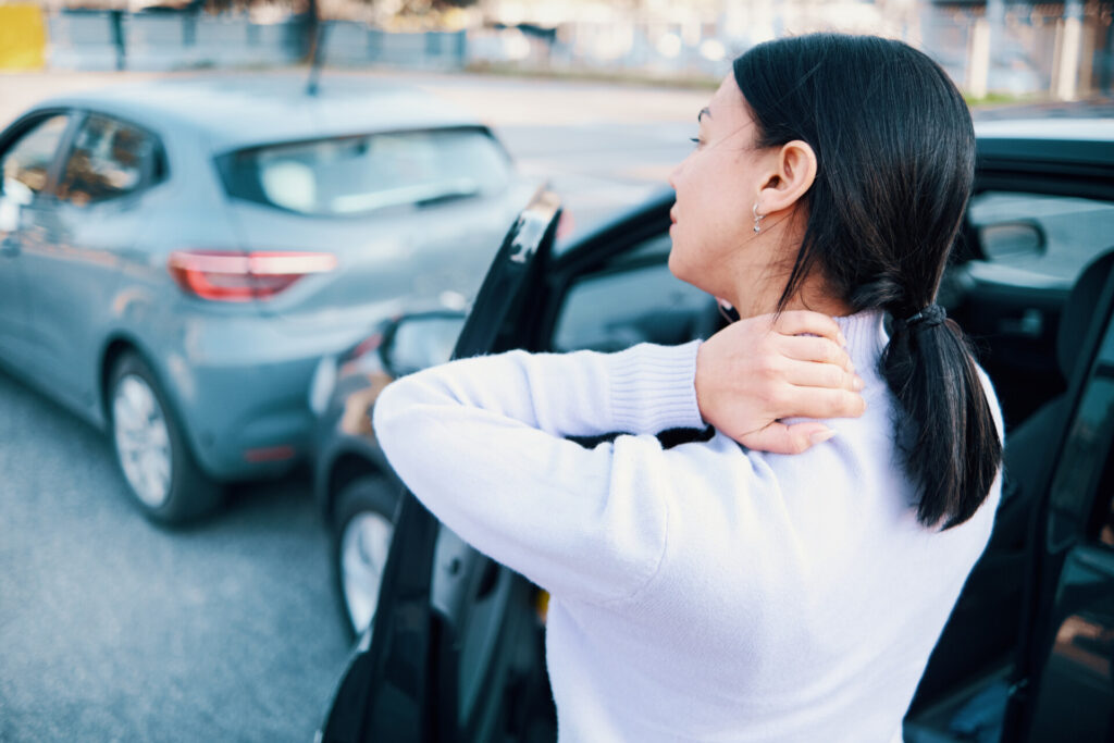 A woman stands by an open car door, holding her neck, with another car parked closely behind her in a parking lot.