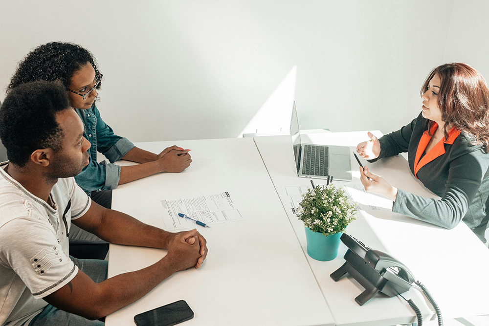 Three people sit at a white table in an office, with two on one side and one on the other, discussing papers and a laptop. A small potted plant and a phone are on the table.