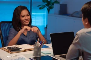 Two people have a conversation at a desk in an office setting. One woman is smiling and holding a pen, while the other person’s back is visible. Laptops and paperwork are on the desk.