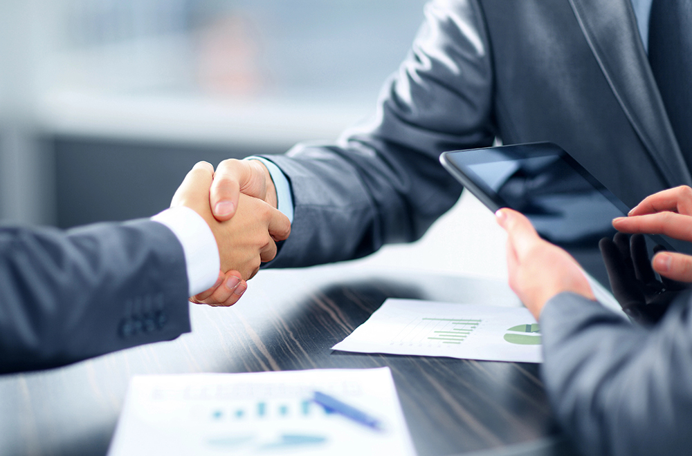 Two people in business suits shaking hands across a table with documents and a tablet, suggesting a business meeting or agreement.