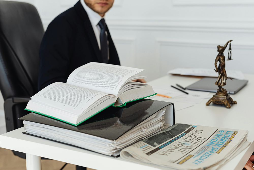A person in a suit sits at a desk with an open book, a stack of files, newspapers, and a small statue of Lady Justice.