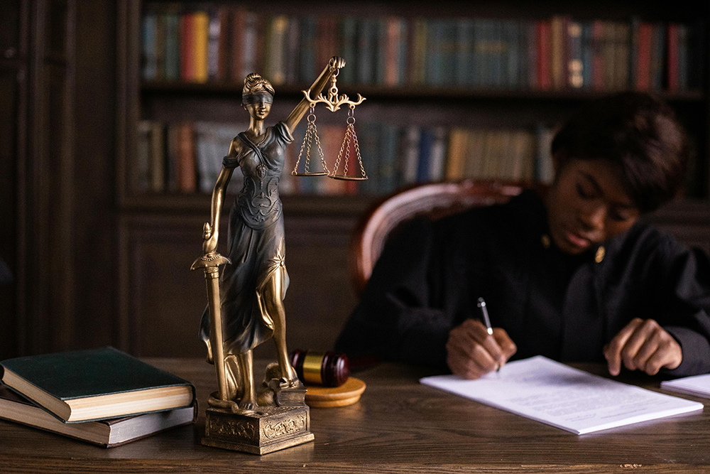 A statue of Lady Justice stands on a desk, with books and a gavel nearby, while a person in judicial robes writes on a document in the background.