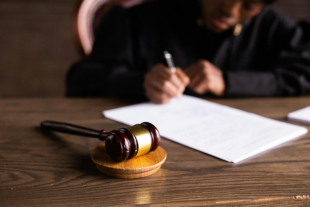 A wooden judge's gavel rests on a desk while a person in a black robe writes on documents in the background.