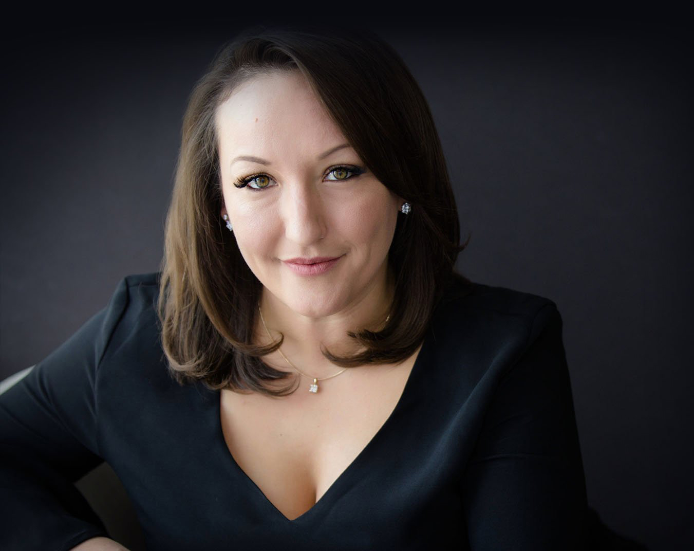 A woman with shoulder-length brown hair, wearing a black top and necklace, poses against a dark background.