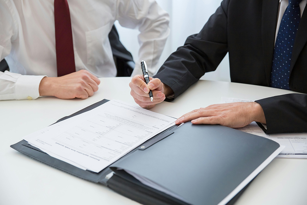 Two people in business attire sit at a table; one holds a pen and appears to be signing a document on a clipboard while the other observes.