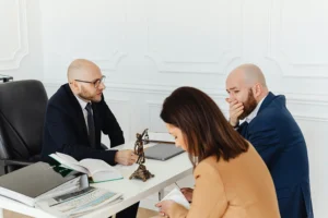Three people in formal attire sit at a desk with legal documents and a statue of Lady Justice, engaged in a serious discussion.