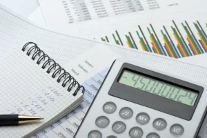 A calculator, pen, and notebook with handwritten numbers rest on financial documents and charts displaying graphs and data.