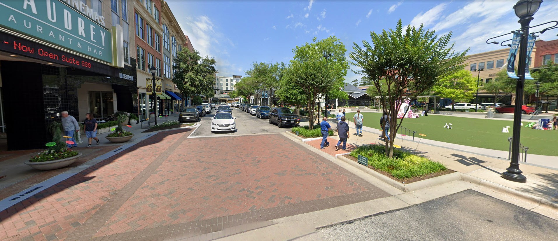 A brick-paved street with parked cars, shops on the left, and people walking near a grassy park area on the right under a partly cloudy sky.