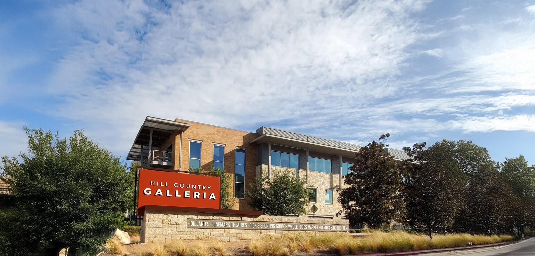 A modern two-story building with a sign reading "Hill Country Galleria" stands under a partly cloudy sky, surrounded by trees and landscaping.