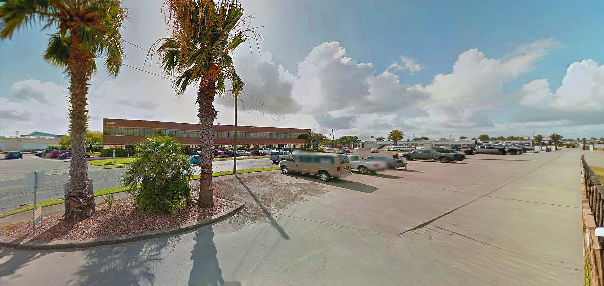 A parking lot with parked cars, palm trees, and a two-story brick building in the background under a partly cloudy sky.