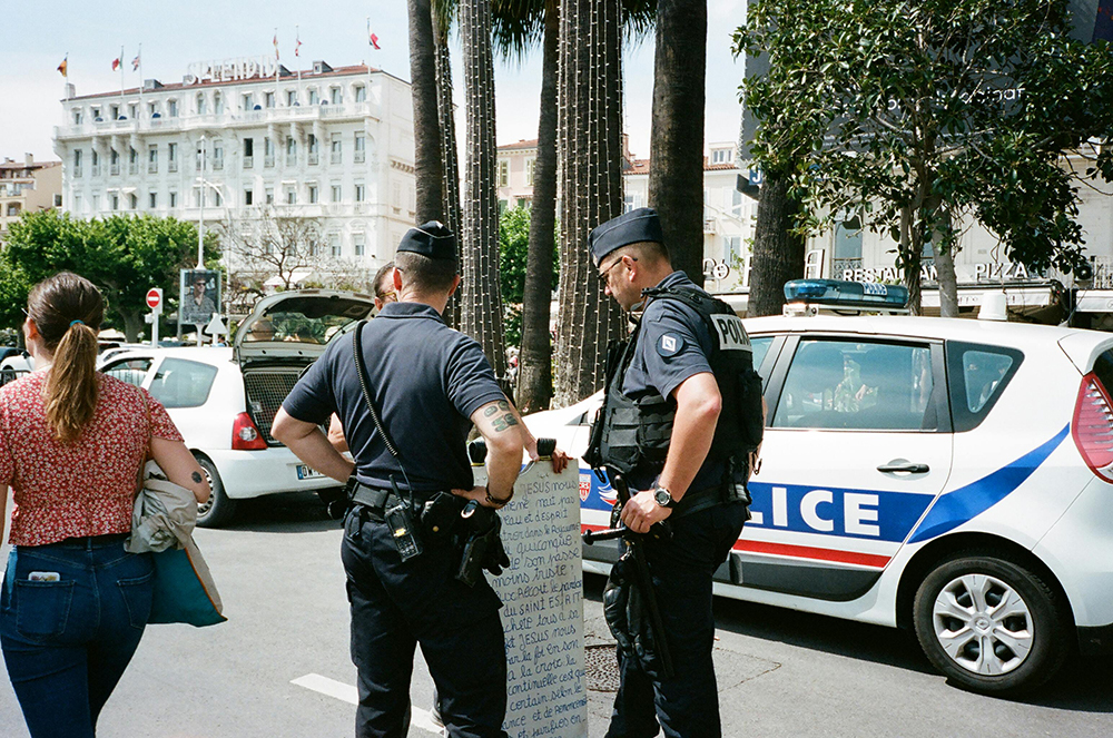 Two police officers stand near police cars on a city street, reading a handwritten sign held by a person. A woman walks away in the foreground. A large hotel is visible in the background.