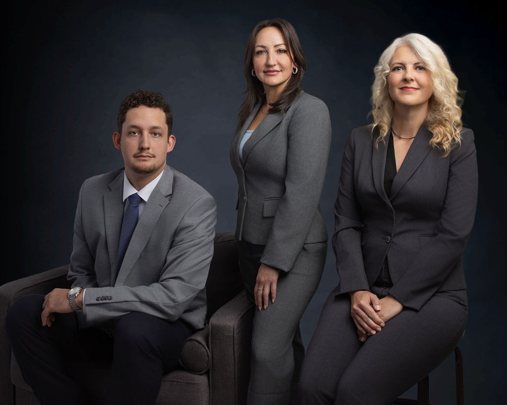Three people in business suits pose for a formal group portrait against a dark background; two women are standing and sitting, and one man is seated on a chair.