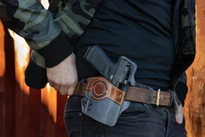 Person wearing camouflage jacket and black shirt, carrying a handgun in a leather holster attached to a brown belt.